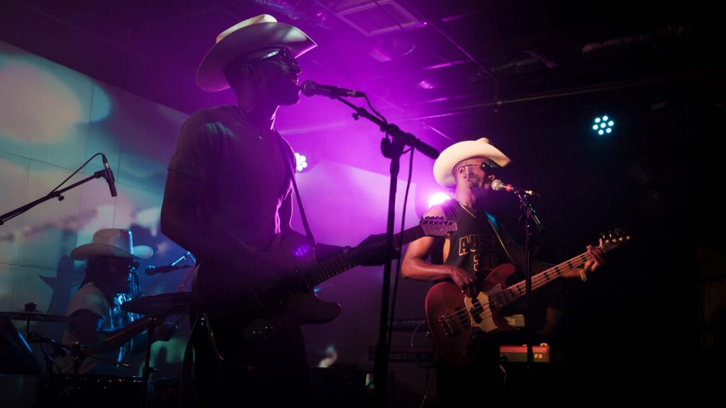 Austin-based musicians The Bros Fresh on stage at Empire Control Room playing guitars wearing cowboy hats on stage backlit with purple light.