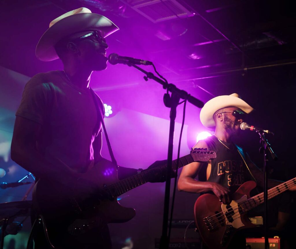 Austin-based musicians The Bros Fresh on stage at Empire Control Room playing guitars wearing cowboy hats on stage backlit with purple light.